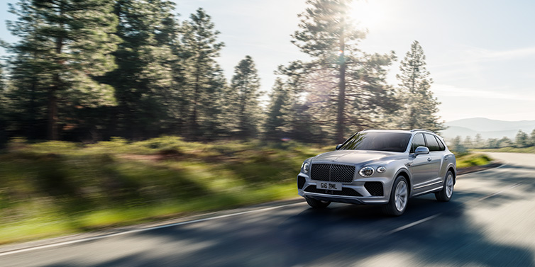 Bentley Monaco Bentley Bentayga SUV front three quarter in Moonbeam silver paint driving dynamically along a tree-lined road