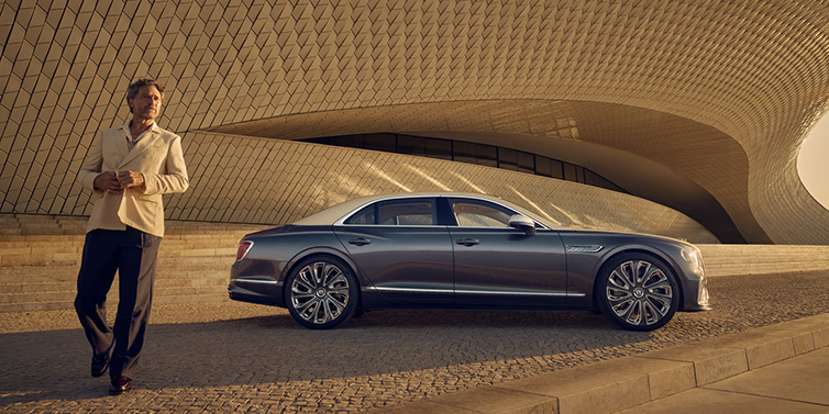 Bentley Monaco Bentley Flying Spur Mulliner sedan in Rose Gold over Magnetic duotone paint parked by a curved wall with person in suit jacket standing on the left